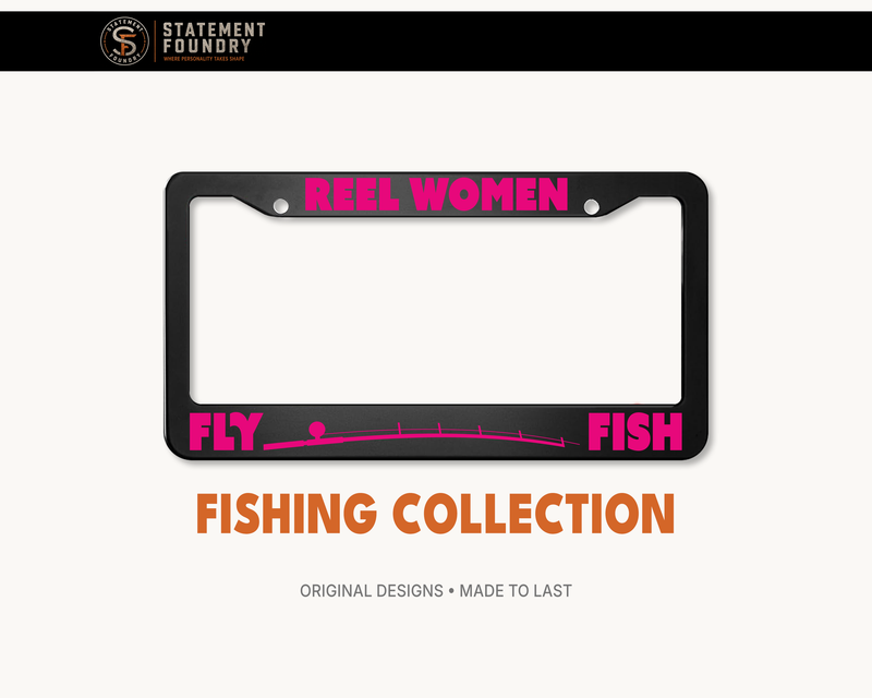 Black metal license plate frame featuring “Reel Women Fly Fish” in bold pink lettering with a fly-fishing graphic.